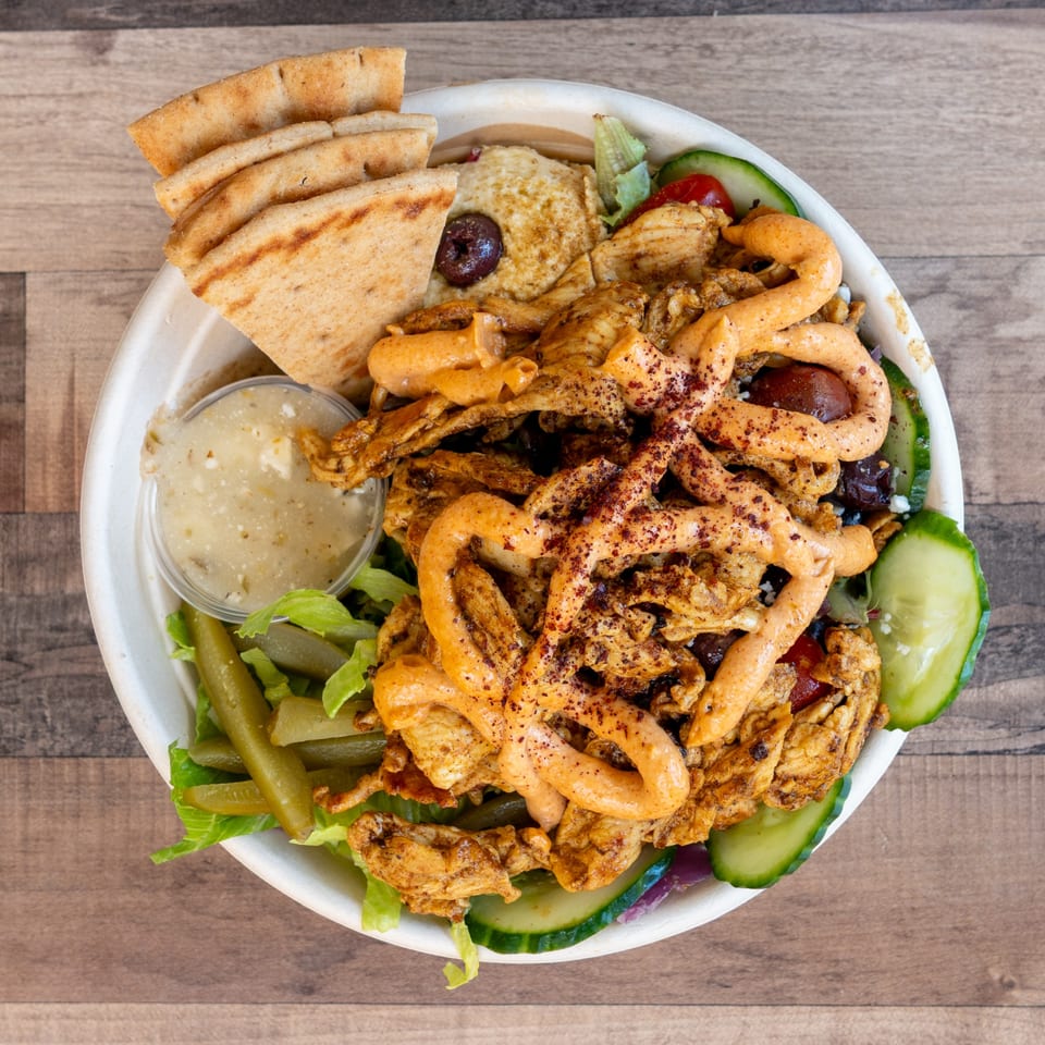 Overhead view of a chicken shawarma bowl with pita, hummus, pickles, and a side of sauce.