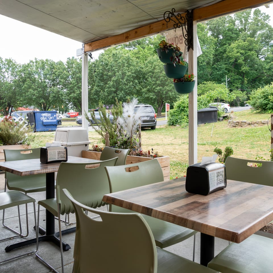 Outdoor patio seating area with light green chairs and wooden tables, surrounded by greenery.