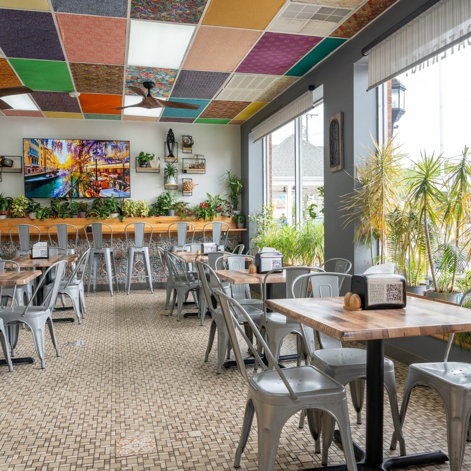 Restaurant interior with colorful ceiling tiles, metal chairs, wooden tables, and plants by large windows.