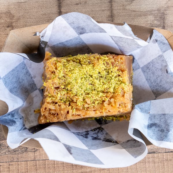 A square piece of baklava with shredded coconut and green pistachio crumbles in a paper-lined box.
