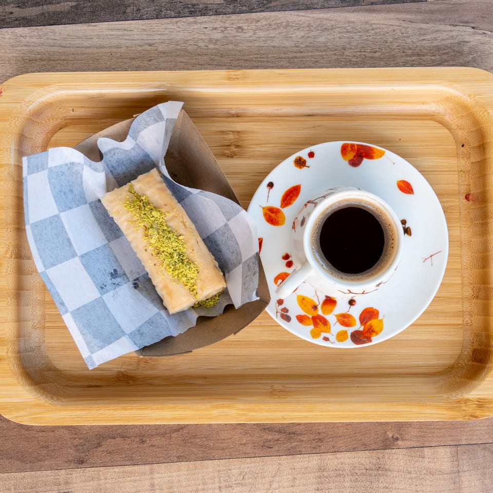 A wooden tray holds a pastry with pistachios in checkered paper and a cup of coffee.