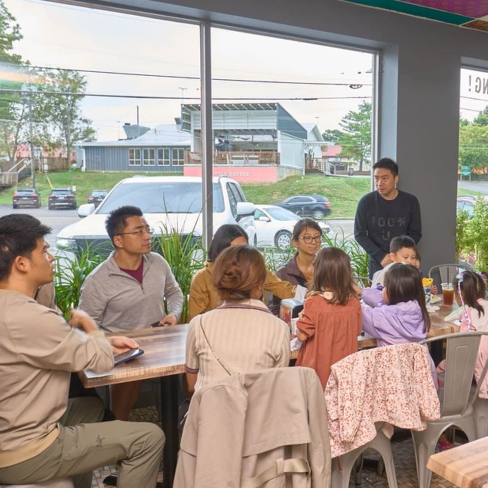 A group of adults and children sitting at tables inside a restaurant with large windows.