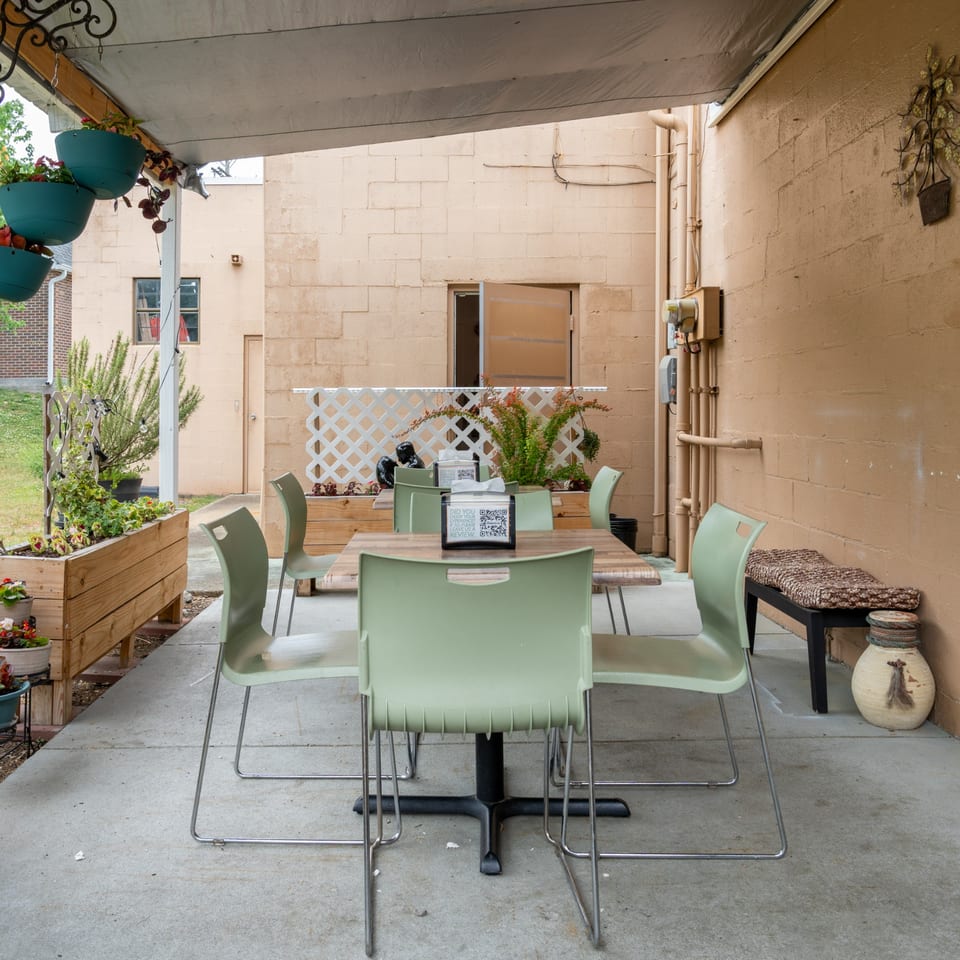 Outdoor patio dining area with light green chairs, wooden tables, and planters under a white awning.