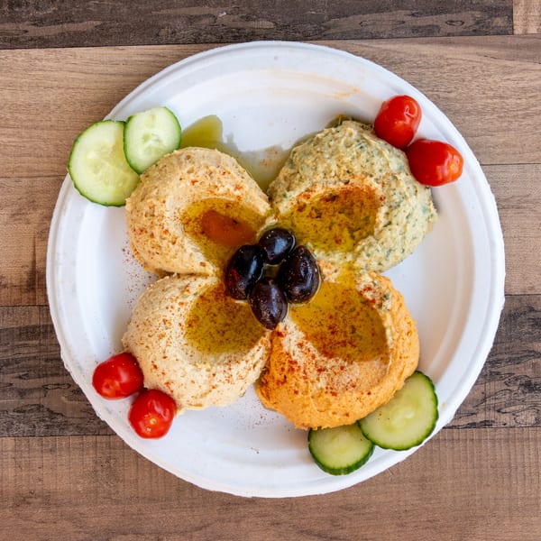 Four different hummus varieties, garnished with olives, cherry tomatoes, and cucumber slices on a white plate.