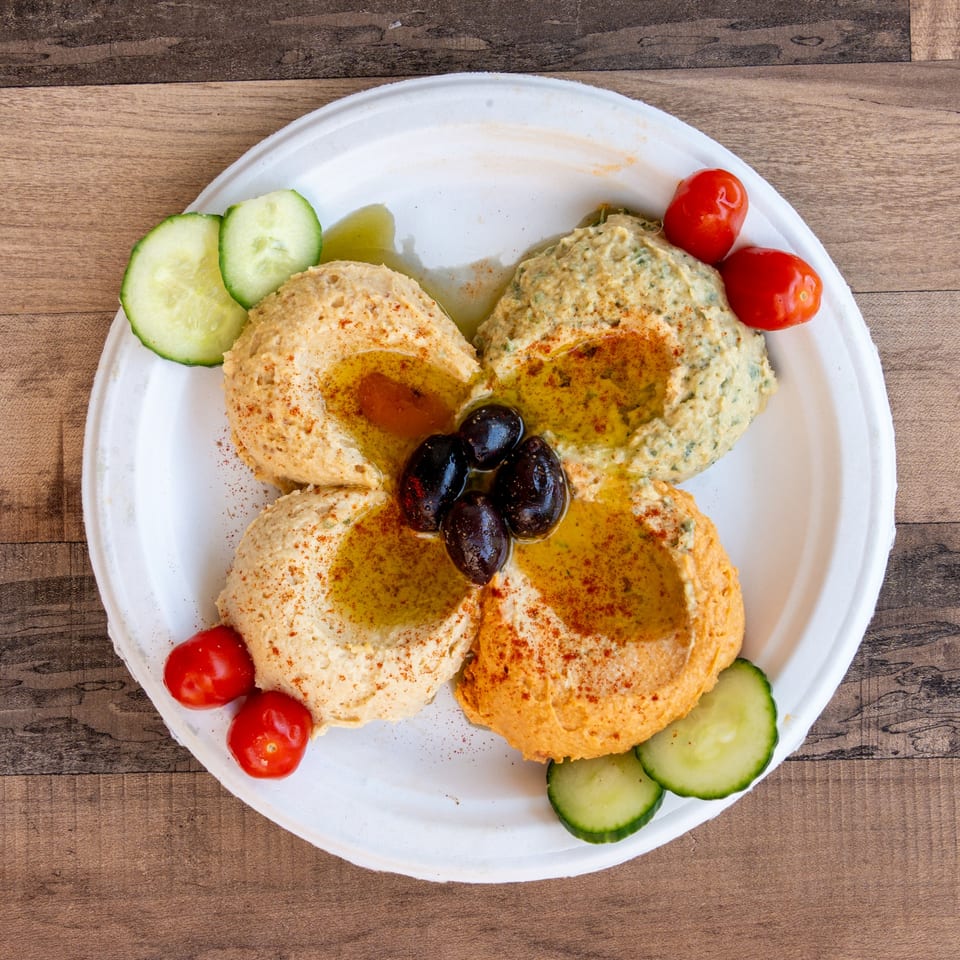 Four different hummus varieties, garnished with olives, cherry tomatoes, and cucumber slices on a white plate.