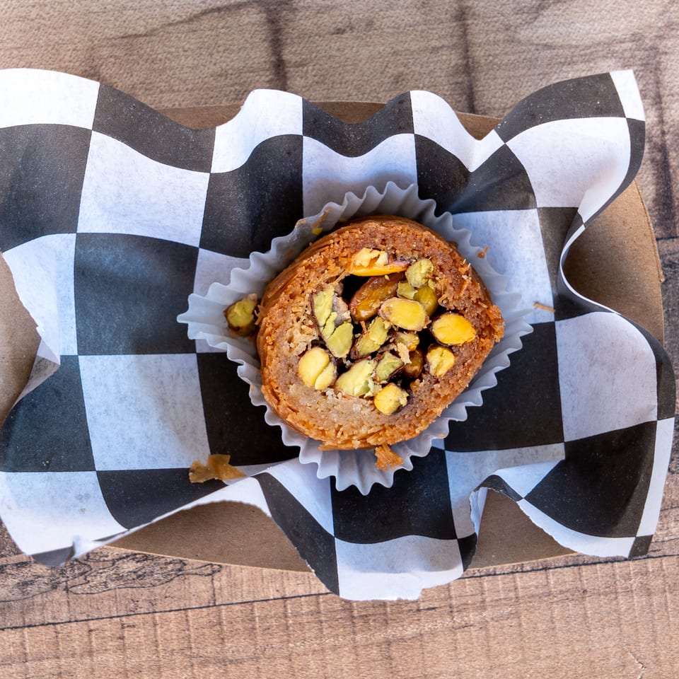 A single baklava pastry, topped with chopped pistachios, in a black and white checkered paper liner.