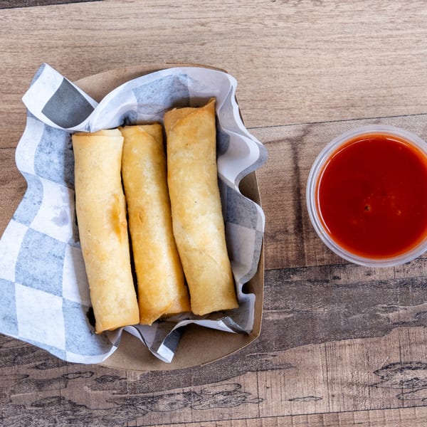 Three golden-brown spring rolls in a checkered paper-lined container with a side of red dipping sauce.