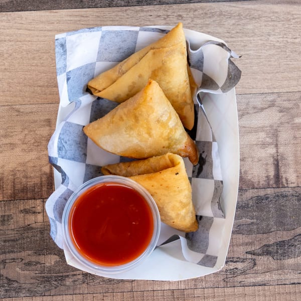 Three golden-brown samosas and a small cup of red dipping sauce in a checkered paper tray.